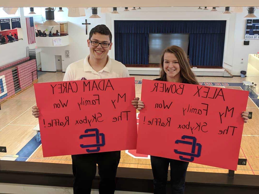Two students in balcony overlooking the gymnasium holding signs announcing themselves as the winners of the Skybox Raffle.
